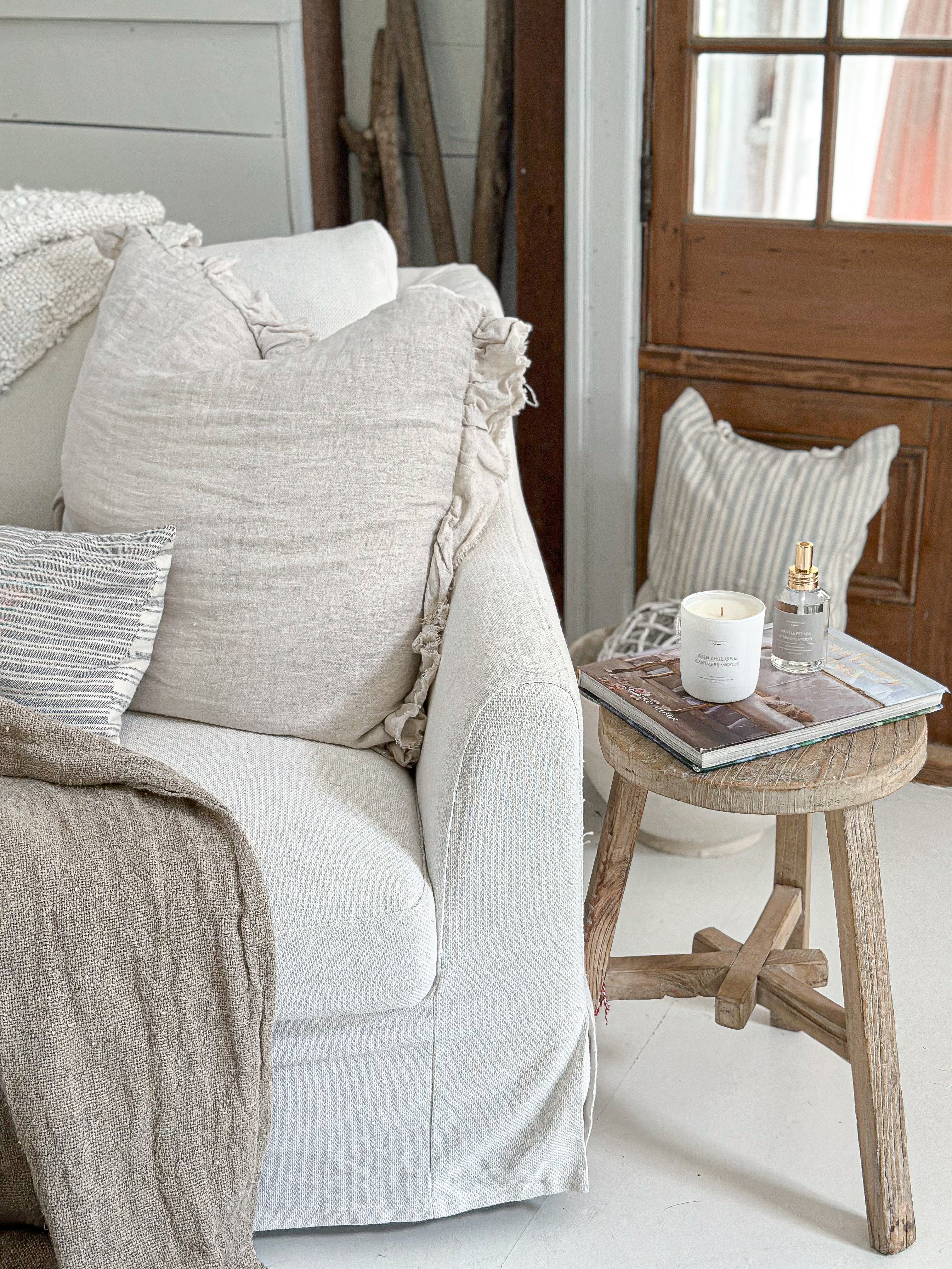 interior home setting with a large white comfy couch, neutral accents and textiles, linen pillows, and a wooden stool with a design book and a candle and room spray displayed on top.