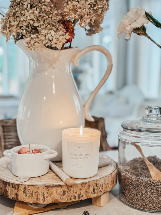 Candle with a Calabrian Lemon & Lilac Leaves Scent on a rustic surface with a pitcher, bowl, and jar in the background.