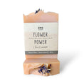 Load image into Gallery viewer, Awe Soap 'Flower Power' Shea Butter Body Bar in a peach-pink color with dried lavender flowers embedded in the bar, shown with its paper label on a white background.
