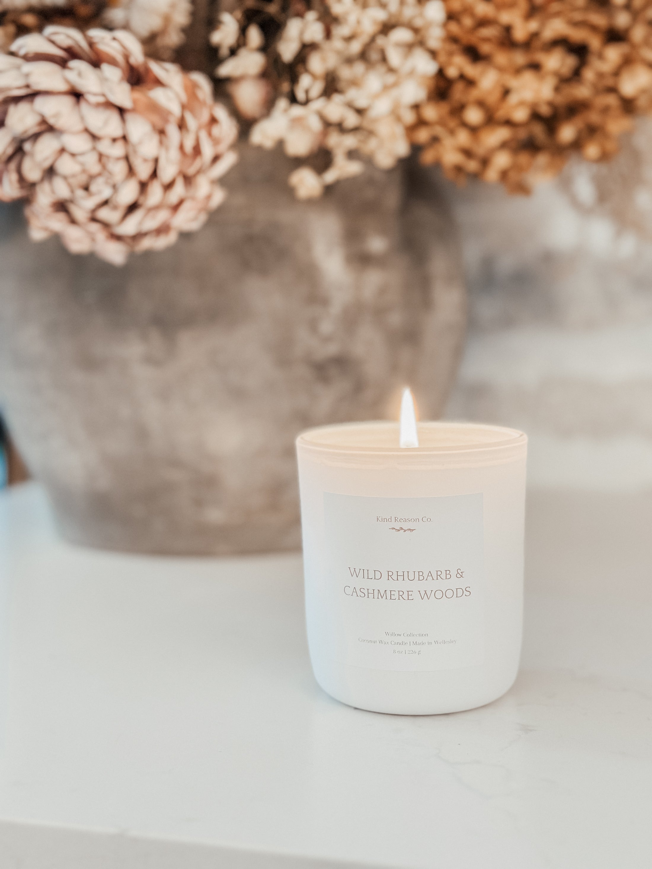 Coconut Wax Candle with 'Wild Rhubarb & Cashmere Wood' scent in front of a vase with dried flowers on a white surface